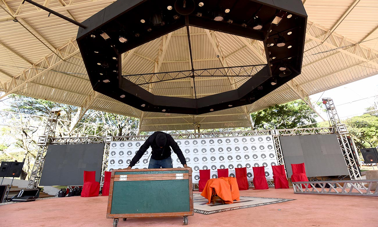 Audiodescrição: no palco de amplo teatro de arena, imagem frontal em plano geral de homem que arrasta com as mãos uma grande caixa de madeira com rodinhas, como se estivesse a organizar o local para um evento. Atrás dele, ao fundo na imagem, cerca de 10 cadeiras dispostas uma ao lado da outra, recobertas com tecido vermelho, e uma ampla estrutura de ferro, disposta na horizontal, com um tipo de banner ao centro com vários logotipos da Unicamp. O teto do local é de estruturas e telhas de metal. Imagem 1 de 1