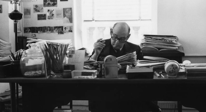 Imagem em preto e branco mostra um homem sentado em uma mesa de trabalho rodeado de livros.
