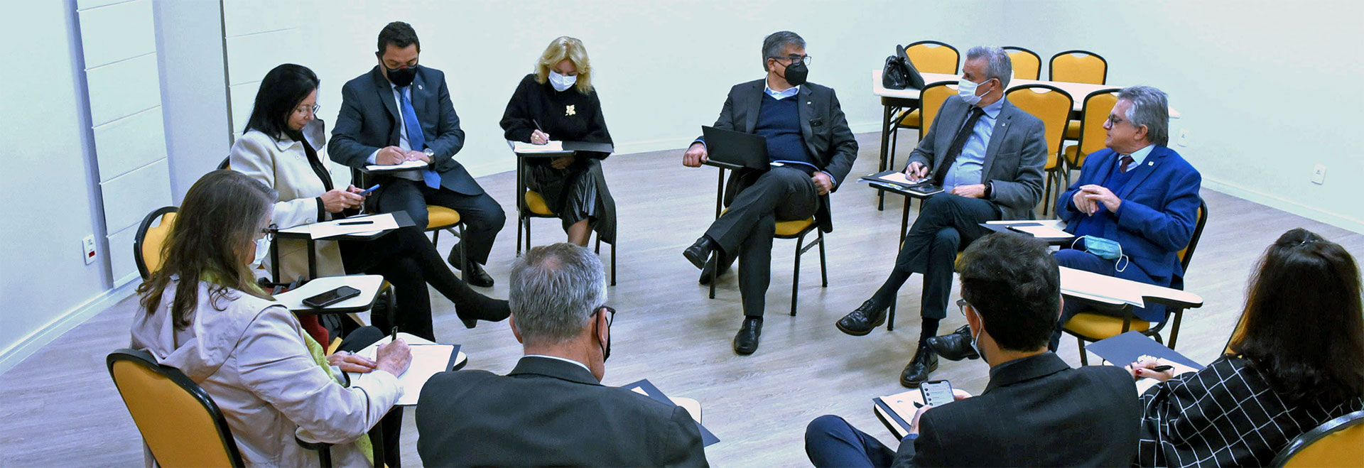 Encontro de 15 de junho definiu como a primeira atividade conjunta do semestre o curso de graduação “Sustentabilidade Empresarial” com início previsto para o dia 17 de agosto Audiodescrição (AD): Em uma ampla sala, imagem em plano geral de 10 pessoas sentadas em carteiras escolares com braço, de maneira a formar uma grande roda. Todos parecem concentrados, sendo que alguns escrevem com caneta em folhas de papel sulfite, outros operam smartphones e conversam. Todos usam máscara de proteção respiratória e vestem traje social. Imagem 1 de 1.