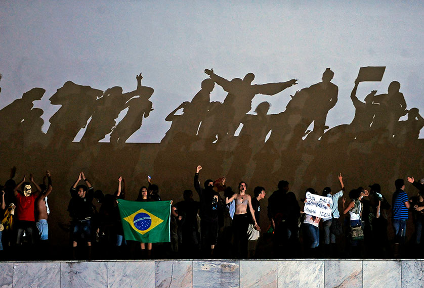 Audiodescrição: imagem colorida; sobre uma mureta, dezenas de manifestantes com suas respectivas sombras projetadas ao fundo em uma parede branca (Foto: Agência Brasil) 