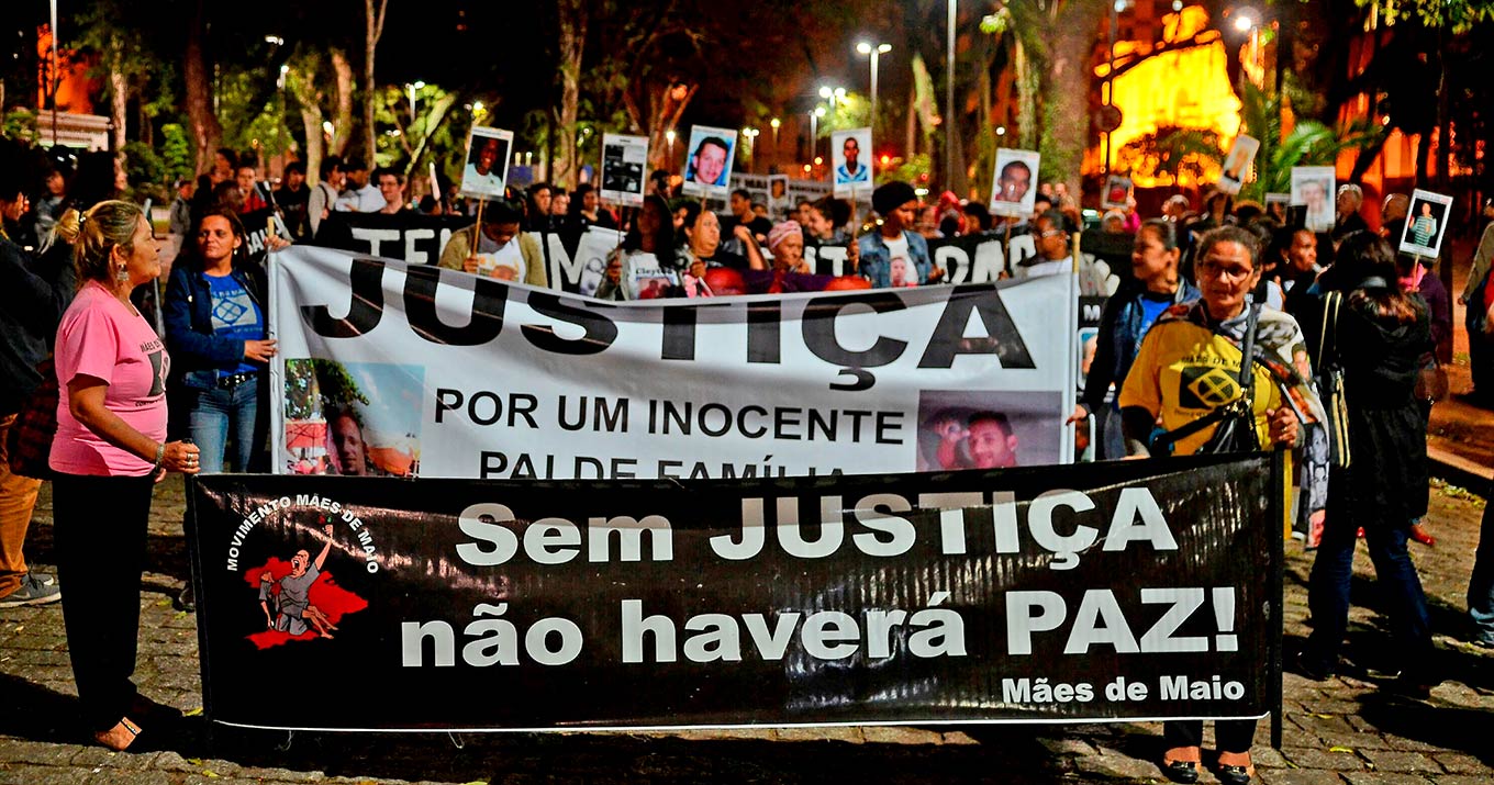 Foto mostra um grupo de mulheres em uma manifestação. Na parte inferior da imagem há um cartaz preto onde está escrito "Sem justiça não haverá paz".