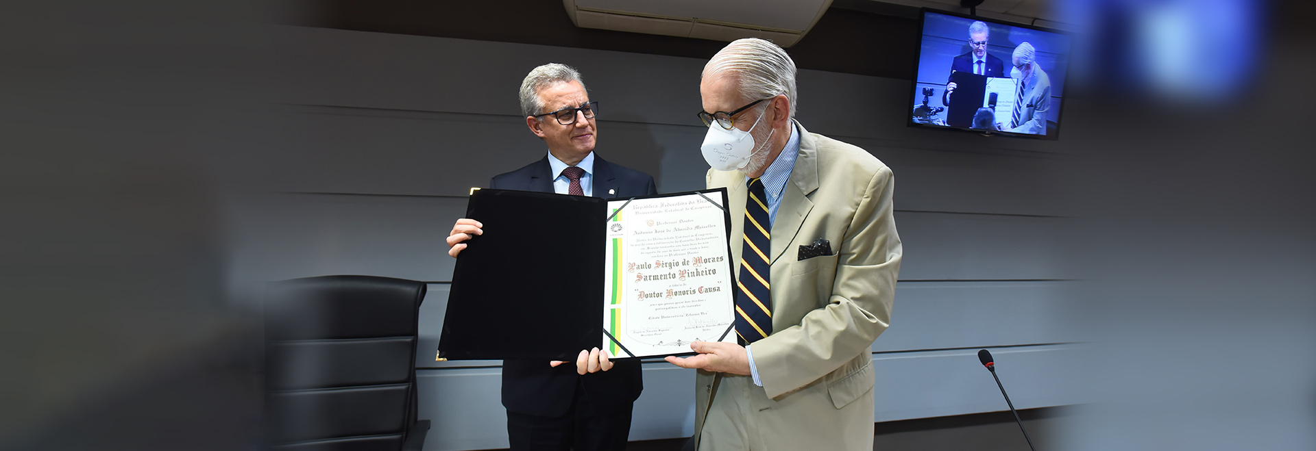 Paulo Sérgio de Moraes Sarmento Pinheiro é o 31º homenageado com o título de Dr. Honoris Causa pela Unicamp Audiodescrição. Foto: Antonio Scarpinetti. Em um auditório, imagem frontal e à curta distância de 2 homens em pé, posicionados um ao lado do outro, sendo que ambos seguram com as mãos uma pasta preta aberta onde há um documento impresso em que se lê em destaque Paulo Sérgio de Moraes Sarmento Pinheiro Doutor Honoris Causa. O homem à direita da imagem olha para o documento, enquanto o outro sorri e o observa. Ambos usam óculos, terno e gravata. Imagem 1 de 1.