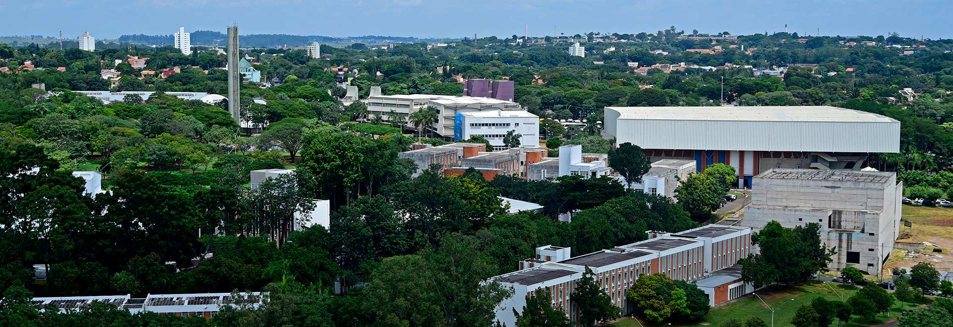 A Unicamp situa-se entre as posições 401 e 500 na lista geral do ranking THE e em terceira colocação na América Latina Audiodescrição. Foto: Antonio Scarpinetti. Imagem panorâmica, obtida de algum ponto mais elevado, de área da Unicamp, onde se destacam várias árvores de grande porte em meio à edificações de concreto, a maioria de poucos andares, que compõem as instalações da universidade e de bairros vizinhos. A imagem se estende por quilômetros, das áreas em primeiro plano até o final do horizonte, delimitado pelo céu azul. Imagem 1 de 1.
