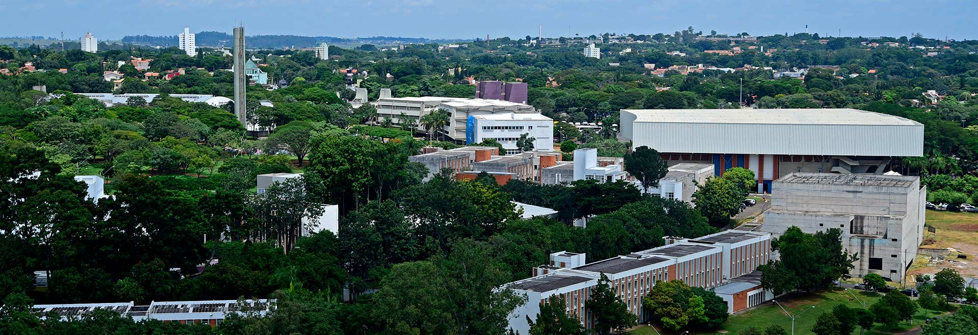 Audiodescrição. Foto: Antonio Scarpinetti. Imagem panorâmica, obtida de algum ponto mais elevado, de área da Unicamp, onde se destacam várias árvores de grande porte em meio à edificações de concreto, a maioria de poucos andares, que compõem as instalações da universidade e de bairros vizinhos. A imagem se estende por quilômetros, das áreas em primeiro plano até o final do horizonte, delimitado pelo céu azul. Imagem 1 de 1.