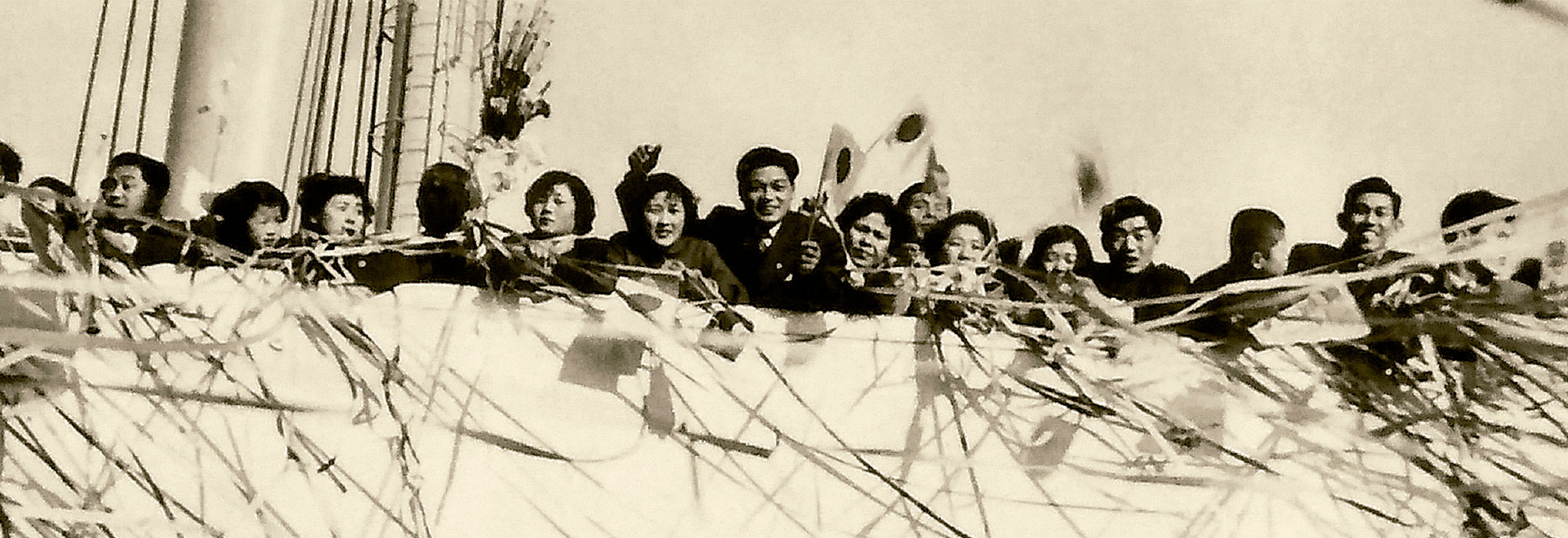Audiodescrição: foto em preto e branco de um grupo de pessoas na borda de um navio. Imagem 1 de 1