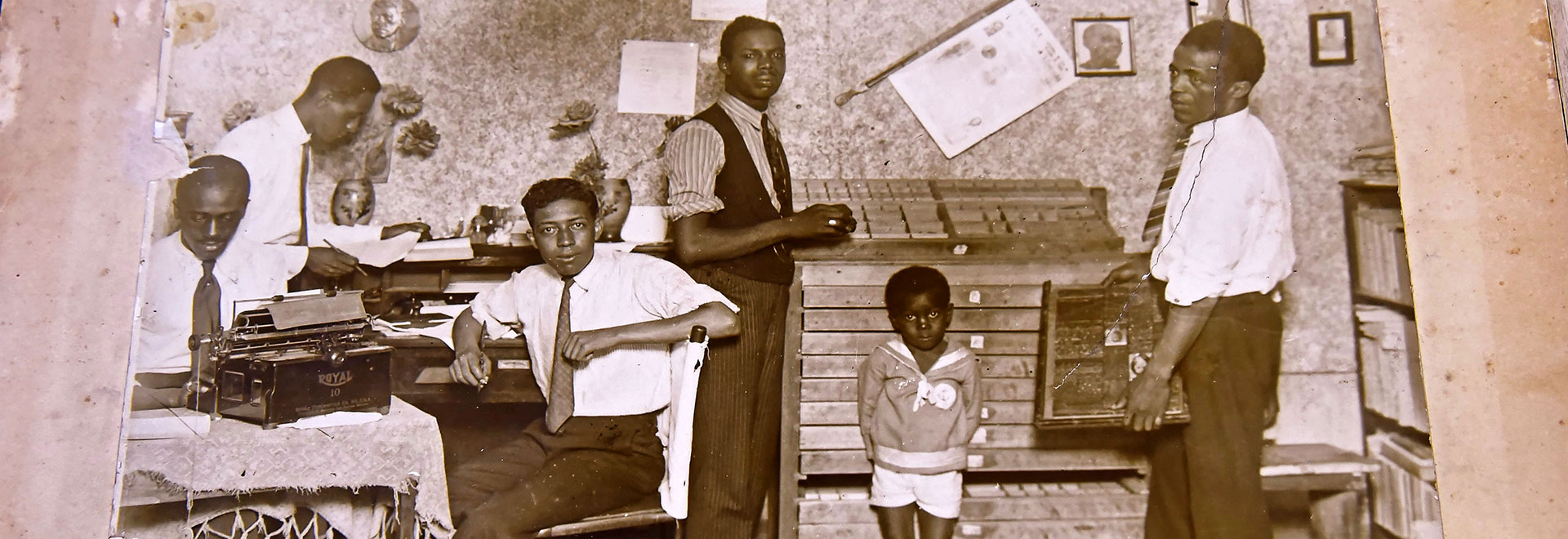 Audiodescrição. Foto antiga em tom sépia, da primeira metade do século 20, de 5 homens e uma criança em uma sala, sendo, a partir da esquerda, homem sentado em cadeira que usa máquina de datilografia apoiada em uma mesa; outro sentado e que segura folha de papel e caneta; outro sentado em cadeira com caneta na mão; um homem em pé; uma criança em pé, e o último homem em pé, que segura uma caixa com as mãos. Ao fundo, parede com quadros e capas de jornal. Os homens usam camisa social e gravata. Imagem 1 de 1