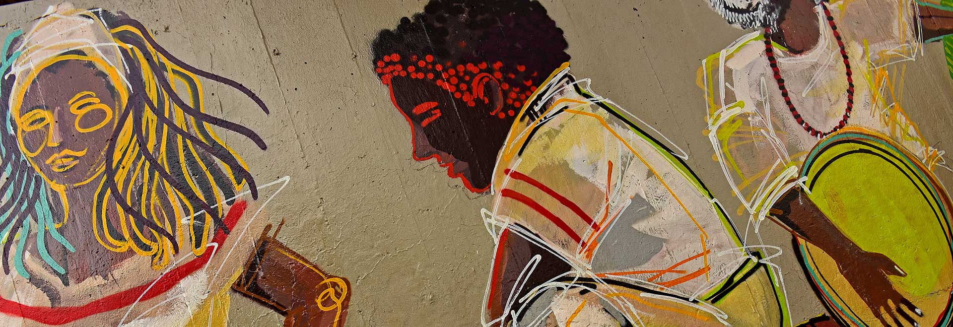Grafite do artista Diogenes Moura alusivo à cultura afro estampado no Teatro de Arena da Unicamp; atividades com Peck e Mbembe integram mês dedicado à Consciência Negra Audiodescrição. Foto: Antonio Scarpinetti. Foto de grafite colorido em muro de concreto que traz 3 pessoas. A primeira, à esquerda, imagem frontal do rosto e busto de mulher com cabelos amarelos e marrons, que usa bandana branca. Ao centro, imagem em perfil de jovem agachado, voltado à esquerda da foto, que usa camisa branca com riscos coloridos. Na última, imagem frontal de pessoa, do pescoço para baixo, que usa roupa branca, colar de contas vermelhas e pretas e toca instrumento de percussão. Imagem 1 de 1
