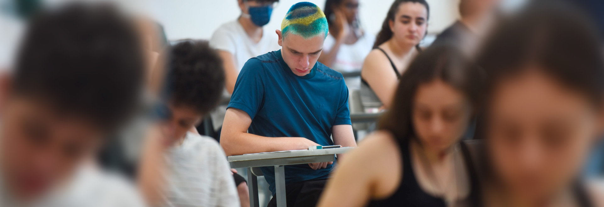 Audiodescrição. Foto: Antonio Scarpinetti. Em uma sala de aula, imagem em perspectiva à média distância de jovem sentado em carteira escolar de braço, voltado para a direita da foto, que realiza prova do vestibular, sendo que ele mantém a cabeça baixada, concentrado, e o braço direito apoiado sobre a carteira, enquanto lê as questões da prova impressa. Ele tem o cabelo curto e com desenho da bandeira do Brasil pintada. Ao redor dele, em imagem desfocada, outras pessoas também fazem a prova. Imagem 1 de 1