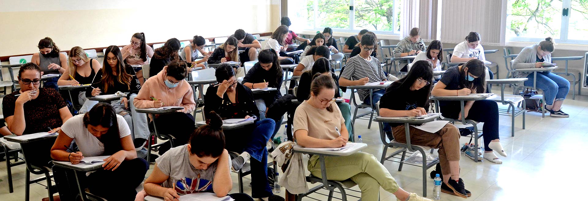 O segundo dia de provas será de conhecimentos específicos, conforme a opção de curso Audiodescrição. Foto: Antoninho Perri. Em ampla sala de aula, imagem em perspectiva à média distância de cerca de 20 pessoas sentadas em carteiras escolares de braço, voltadas para a direita da imagem, que realizam prova. A maioria mantêm a cabeça baixada e escreve com caneta nas folhas de papel das prova que estão apoiadas sobre as carteiras. As carteiras estão posicionadas uma ao lado da outra, formado várias fileiras horizontais. Ao fundo, ampla janela de vidro e persianas verticais. Imagem 1 de 1