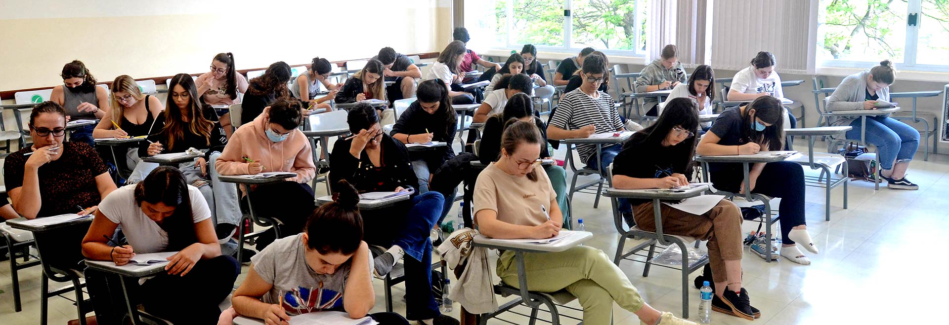 Audiodescrição. Foto: Antoninho Perri. Em ampla sala de aula, imagem em perspectiva à média distância de cerca de 20 pessoas sentadas em carteiras escolares de braço, voltadas para a direita da imagem, que realizam prova. A maioria mantêm a cabeça baixada e escreve com caneta nas folhas de papel das prova que estão apoiadas sobre as carteiras. As carteiras estão posicionadas uma ao lado da outra, formado várias fileiras horizontais. Ao fundo, ampla janela de vidro e persianas verticais. Imagem 1 de 1