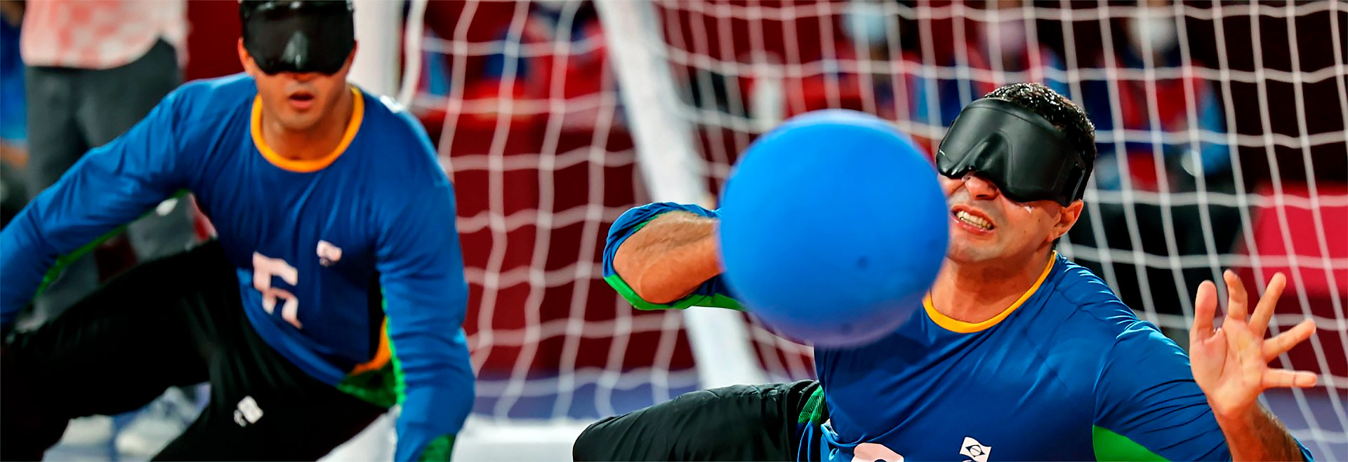 Audiodescrição: em uma quadra de esportes, imagem frontal à curta distância de homem deitado lateralmente no chão, à direita da foto, durante partida de goalball. Ele rebate com a mão direita uma bola azul, com a expressão de quem faz força mantendo os dentes cerrados. Ele veste camisa azul de mangas compridas e tem olhos vendados. À esquerda da foto, ao lado dele, outro atleta participa do jogo, também com camisa azul e olhos vendados. Ao fundo, imagem desfocada das balizas e redes do gol. Imagem 1 de 1