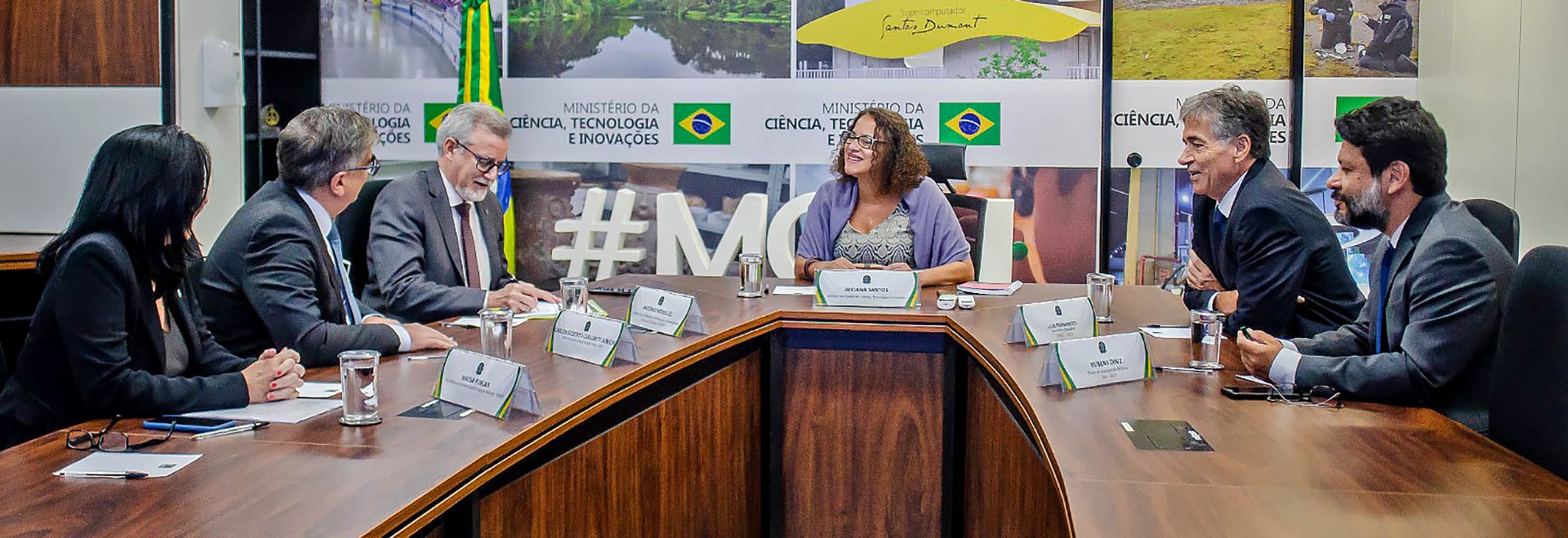 Audiodescrição. Em sala de reuniões, imagem frontal à média distância de mulher sentada em cadeira, voltada à esquerda da foto, que sorri com braços sobre moderna e ampla mesa de madeira oval. À esquerda e à direita dela, 5 pessoas sentadas em cadeiras e com braços sobre a mesa. Na mesa há copos de vidro com água e placas com identificação de nome. Todos usam roupa social. Ao fundo, amplo painel com 4 fotos coloridas, bandeiras do Brasil, e o nome Ministério da Ciência, Tecnologia e Inovações. Imagem 1 de 1