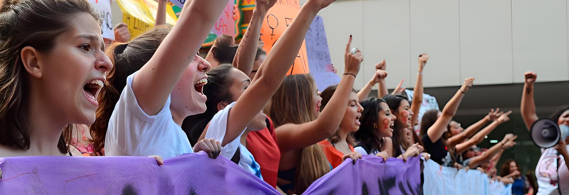 Manifestação contra feminicídio (Ni Una a Menos) na Avenida Paulista; pesquisa referente a 2021 constata que 76,5% dos casos ocorrem em casa Audiodescrição. Foto: Rovena Rosa. Em área externa, imagem em perspectiva à média distância de cerca de 20 mulheres em pé, voltadas para a direita da foto e posicionadas uma ao lado da outra, durante manifestação. Elas falam com vigor e a maioria mantém o braço direito erguido, com punho cerrado, enquanto seguram com a outra mão extensas faixas horizontais na altura do peito. Atrás delas, outras pessoas seguram cartazes coloridos, erguendo-os. Algumas têm pintado no rosto o símbolo feminino Imagem 1 de 1