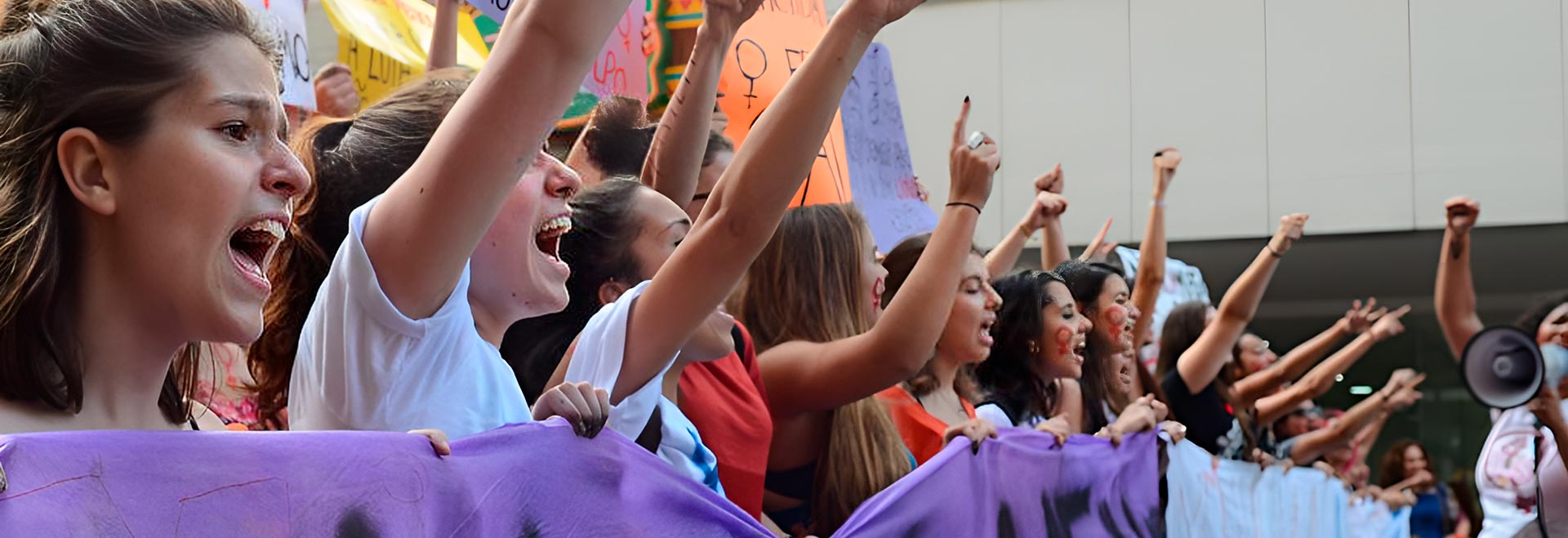 Audiodescrição. Foto: Rovena Rosa. Em área externa, imagem em perspectiva à média distância de cerca de 20 mulheres em pé, voltadas para a direita da foto e posicionadas uma ao lado da outra, durante manifestação. Elas falam com vigor e a maioria mantém o braço direito erguido, com punho cerrado, enquanto seguram com a outra mão extensas faixas horizontais na altura do peito. Atrás delas, outras pessoas seguram cartazes coloridos, erguendo-os. Algumas têm pintado no rosto o símbolo feminino  Imagem 1 de 1