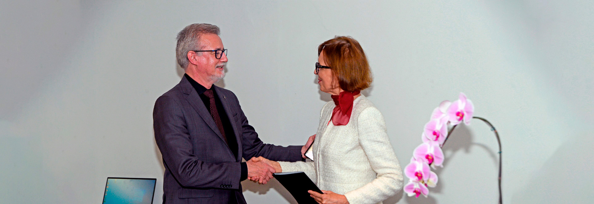 Parceria permitirá a construção de uma usina de geração de energia elétrica por placas fotovoltaicas, a reforma de laboratórios de pesquisa e a contratação de 33 bolsistas de doutorado, pós-doutorado e iniciação científica Audiodescrição. Foto: Antonio Scarpinetti. Imagem em perfil a curta distância de um homem, à esquerda da foto, e uma mulher, ambos em pé, um em frente ao outro, sendo que eles se cumprimentam com a perto de mãos e sorriem discretamente. Ele encosta a mão esquerda no braço direito da mulher, enquanto ela segura uma pasta preta na mão esquerda. Eles usam óculos e vestem roupa social, e ela tem um lenço vermelho no pescoço. À direita da foto, em primeira plano, uma orquídea lilás e branca. Imagem 1 de 1