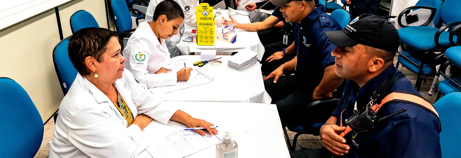O Projeto Integração foi concebido para promover a inserção do terceirizado na comunidade acadêmica Audiodescrição. Foto: Felipe Bezerra. Em área interna, imagem em perfil a média distância de 4 pessoas sentadas em cadeiras, sendo 2 mulheres à esquerda e 2 homens à direita, de frente uns aos outros. Entre eles há mesas quadradas forradas com tecido branco. As mulheres vestem jalecos brancos de mangas compridas, enquanto ele usam bonés e uniforme com camisa azul escura. Eles conversam. Uma mulher escreve com caneta em uma ficha, e, ao fundo uma pessoa faz teste de glicemia no homem à direita. Imagem 1 de 1