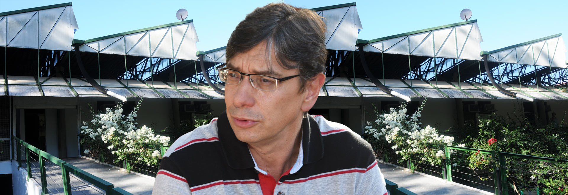 O professor colaborador do Instituto de Economia Márcio Pochmann Audiodescrição. Foto: Antonio Scarpinetti. Montagem com 2 fotos. Em primeiro plano, ao centro, imagem de busto um homem, voltado para a esquerda da foto, sendo que ele usa óculos e traja camisa polo com listras verticais em preto, cinza e linhas vermelhas, intecaladas. Na outra foto, ao fundo de toda imagem, telhado de uma ampla edificação térrea formada por estruturas metálicas do tipo zigue-zague. À frente da edificação há vários arbustos floridos em branco e vermelho. Imagem 1 de 1
