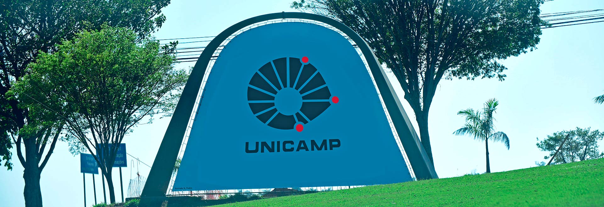 A Reitoria da Unicamp, em complemento às informações apresentadas no comunicado anterior, reitera compromissos Audiodescrição. Foto: Antonio Scarpinetti. Em ampla praça gramada, imagem frontal a longa distância de um banner com cerca de 6 metros de largura por 4 de altura, em formato de arco arredondado, que se encontra afixado em uma estrutura de concreto em formato de arco. O banner traz o logotipo da Unicamp e, abaixo, o nome Unicamp, em letras pretas sobre fundo branco. À direita e à esquerda há algumas árvores de médio e grande portes. O dia está bastante ensolarado. Imagem 1 de 1
