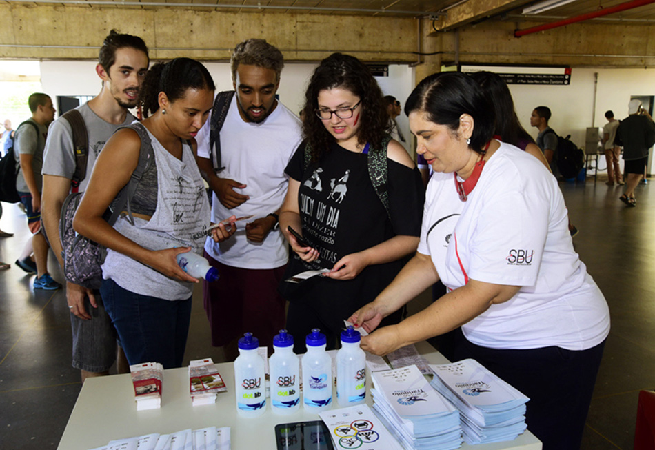 Alunos recebem squeezes durante recepção dos calouros Unicamp 2017 Em um amplo vão de edificação em concreto, quatro jovens, entre homens e mulheres, em pé, um ao lado do outro, observam uma mesa de madeira que está diante deles e onde estão dispostos quatro squeezes, duas pilhas com marcadores de livros, e quatro pilhas com folderes sobre temas como Trânsito e Campus Tranquilo. À direita, uma mulher, funcionária da Unicamp, faz a entrega do material e dá orientações