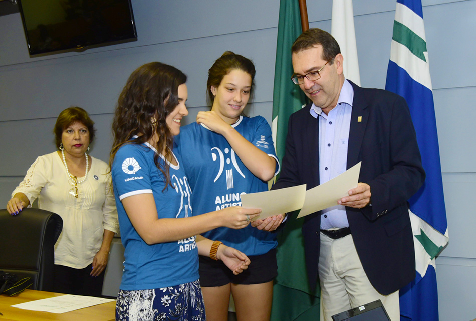 Professor Alvaro Crósta, coordenador geral da Universidade, entrega certificados a alunas Em auditório, duas mulheres, em pé, posicionadas uma ao lado da outra, recebem em mãos, sorrindo, certificados impressos em papel de homem que está também de pé à direita. Ao fundo, à direita, há três bandeiras estendidas em pequenos mastros e um monitor de TV ao alto, sendo que todos são observados por uma mulher que está à esquerda e em pé
