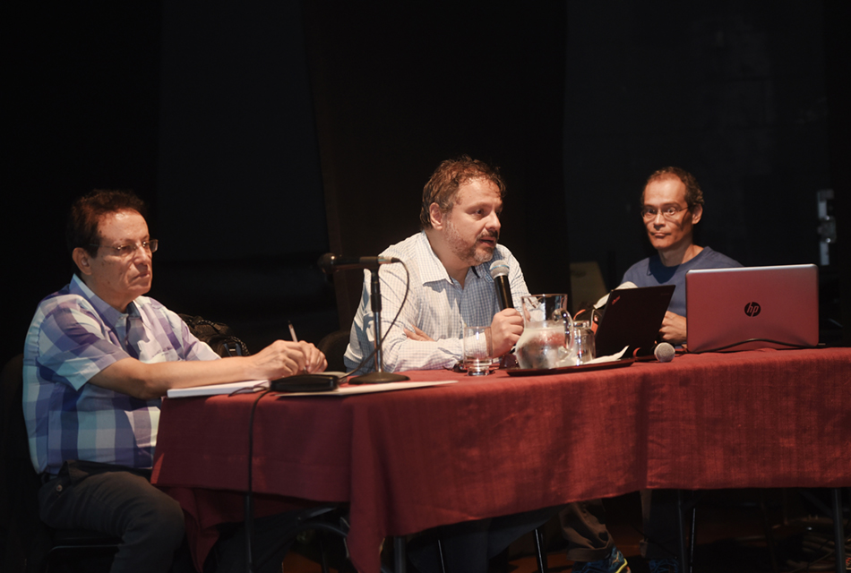 Primeira mesa redonda do dia do Seminário Internacional Música Popular e Política No palco de um anfiteatro, em perspectiva, três homens sentados em cadeiras, um ao lado do outro, sendo que à frente deles há uma extensa mesa retangular de madeira coberta com pano em tom vermelho, sobre a qual apoiam os braços. O homem ao centro fala ao microfone sem fio, segurando-o com a mão direita, sendo que sobre a mesa ainda há dois microfones com fio, um deles fixado em pedestal, e à direita, um notebook aberto