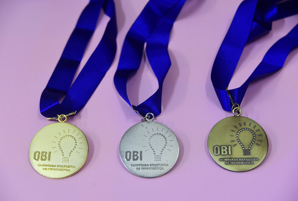 Três medalhas, sendo uma de ouro, uma de prata e outro de bronze, dispostas sobre uma superfície lisa e na cor rosa, uma ao lado da outro, sendo que estão afixadas em um cordão azul. Nas medalhas, há a imagem de uma lâmpada, à direita, e se lê em destaque OBI. Imagem 6 de 6