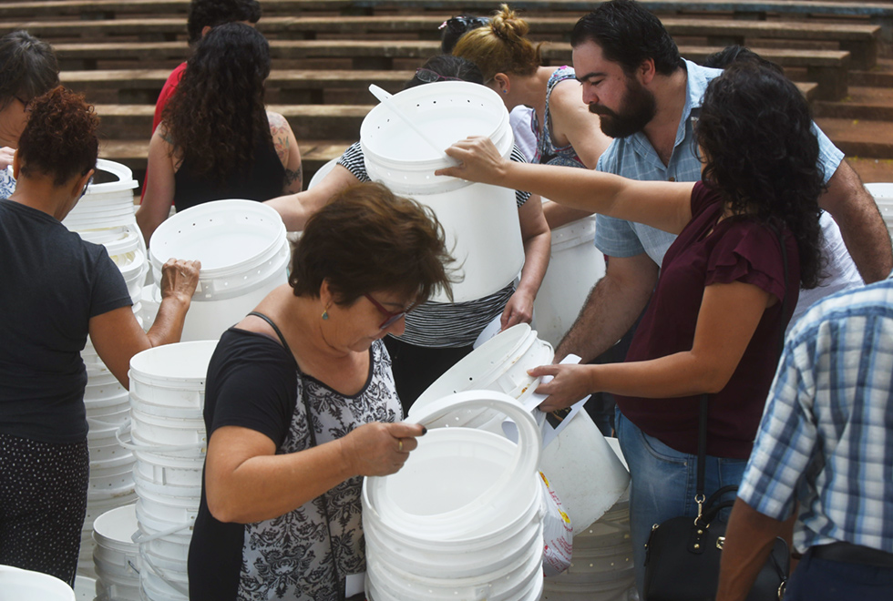 Num ambiente externo, um grupo de onze pessoas manipula baldes de plástico branco e observa seus interiores. Ao fundo, se vê uma arquibancada de concreto. Imagem 13 de 14