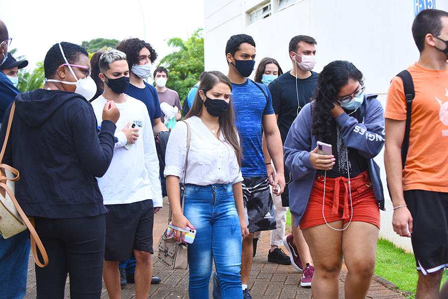 Chegada de candidatos para provas da segunda fase do Vestibular Unicamp 2022, na PUC-Campinas Audiodescrição: Em área externa, imagem em perspectiva e de corpo inteiro, grupo de cerca de 15 pessoas caminha em área com piso intertravado de concreto, da esquerda para a direita na imagem. A maioria veste roupas casuais, com bermuda e tênis. Todos usam máscara de proteção contra covid-19. Ao fundo, à direita na imagem, grande e alta parede de uma edificação. Imagem 2 de 20.
