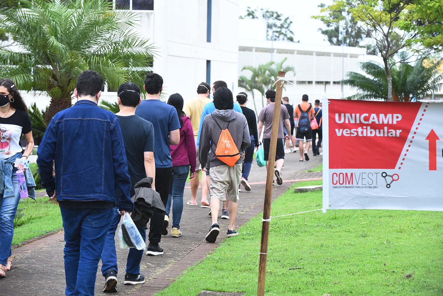 Chegada de candidatos para provas da segunda fase do Vestibular Unicamp 2022, na PUC-Campinas Audiodescrição: Em área externa, imagem em perspectiva e de corpo inteiro, cerca de 15 pessoas, de costas na imagem, caminham formando fila sobre calçamento de concreto, com área gramada à direita e à esquerda, com árvores e palmeiras plantadas. À direita na imagem, parte de uma faixa informativa, onde se lê Unicamp, Vestibular e Comvest. Ao fundo de toda a imagem, parte de uma grande edificação com janelas de vidro.. Imagem 3 de 20.