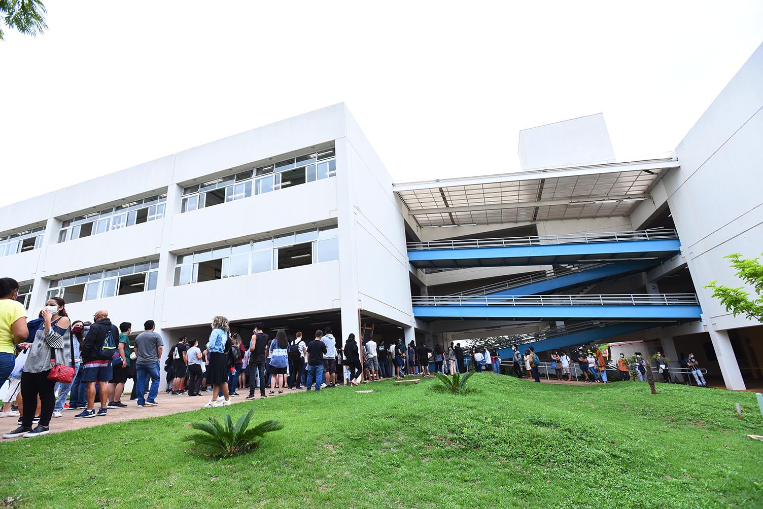 Chegada de candidatos para provas da segunda fase do Vestibular Unicamp 2022, na PUC-Campinas Audiodescrição: Em área externa, imagem panorâmica de baixo para cima, grupo com cerca de 50 pessoas paradas em pé, à frente de uma edificação de dois andares interligada a outro prédio, à direita na imagem, por rampas de ferro pintadas em azul, e com corrimão de proteção. Junto a eles, em primeiro plano na imagem, ampla área gramada. O tempo está nublado. Imagem 4 de 20.
