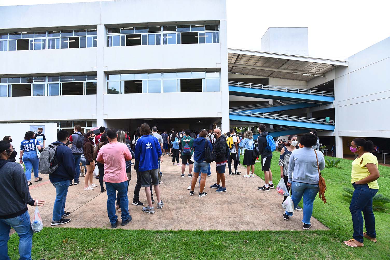 Chegada de candidatos para provas da segunda fase do Vestibular Unicamp 2022, na PUC-Campinas Audiodescrição: Em área externa, imagem panorâmica de grupo com cerca de 50 pessoas paradas em pé sobre ampla área cimentada, à frente de uma edificação de dois andares, com janelas de vidro e interligada a outro prédio, à direita na imagem, por rampas de ferro pintadas em azul, e com corrimão de proteção. Junto a eles, no entorno da área cimentada, ampla área gramada. O tempo está nublado. Imagem 5 de 20.