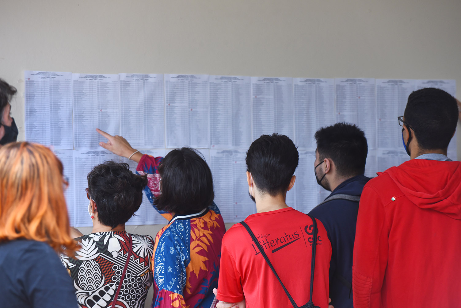 Entrada de candidatos para provas da segunda fase do Vestibular Unicamp 2022, na PUC-Campinas Audiodescrição: Em área interna, imagem à média distância de grupo de 7 pessoas, em pé, de costas na imagem, e dispostas uma ao lado da outra, que observam listagens de nomes impressas em 20 folhas de papel sulfite afixadas em uma parede. Uma mulher, ao centro na imagem, aponta com o dedo indicador direito para um nome na lista. Imagem 8 de 20.