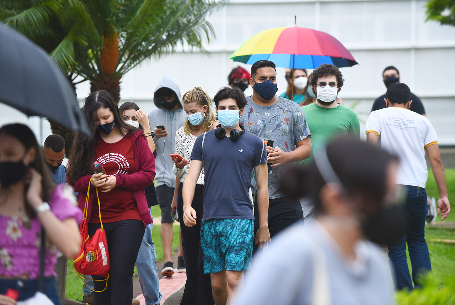 Chegada de candidatos para provas da segunda fase do Vestibular Unicamp 2022, na PUC-Campinas Audiodescrição: Em área externa, imagem de corpo inteiro de cerca de 40 pessoas que caminham em fila, do fundo da imagem para a frente. Há área gramada e árvores no entorno deles. Ao fundo, parte de uma edificação. Todos usam máscaras e vestem roupas casuais como bermuda e camiseta. Destaca-se, ao centro na imagem, um guarda-chuva com as cores do arco-íris. O dia está nublado. Imagem 10 de 20.