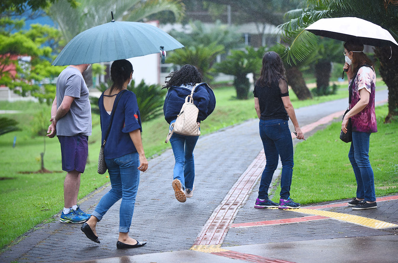 Chegada de candidatos para provas da segunda fase do Vestibular Unicamp 2022, na PUC-Campinas Audiodescrição: Em área externa, imagem de corpo inteiro, grupo de 5 pessoas, em pé, de costas na imagem e dispostas próximas uma da outra. 4 delas estão paradas e observam uma jovem que corre, ao centro da imagem. Todos estão parados em área com piso intertravado que forma caminhos em ampla área gramada e com várias árvores no entorno. Duas pessoas seguram guarda-chuvas abertos. O piso está molhado.. Imagem 11 de 20.