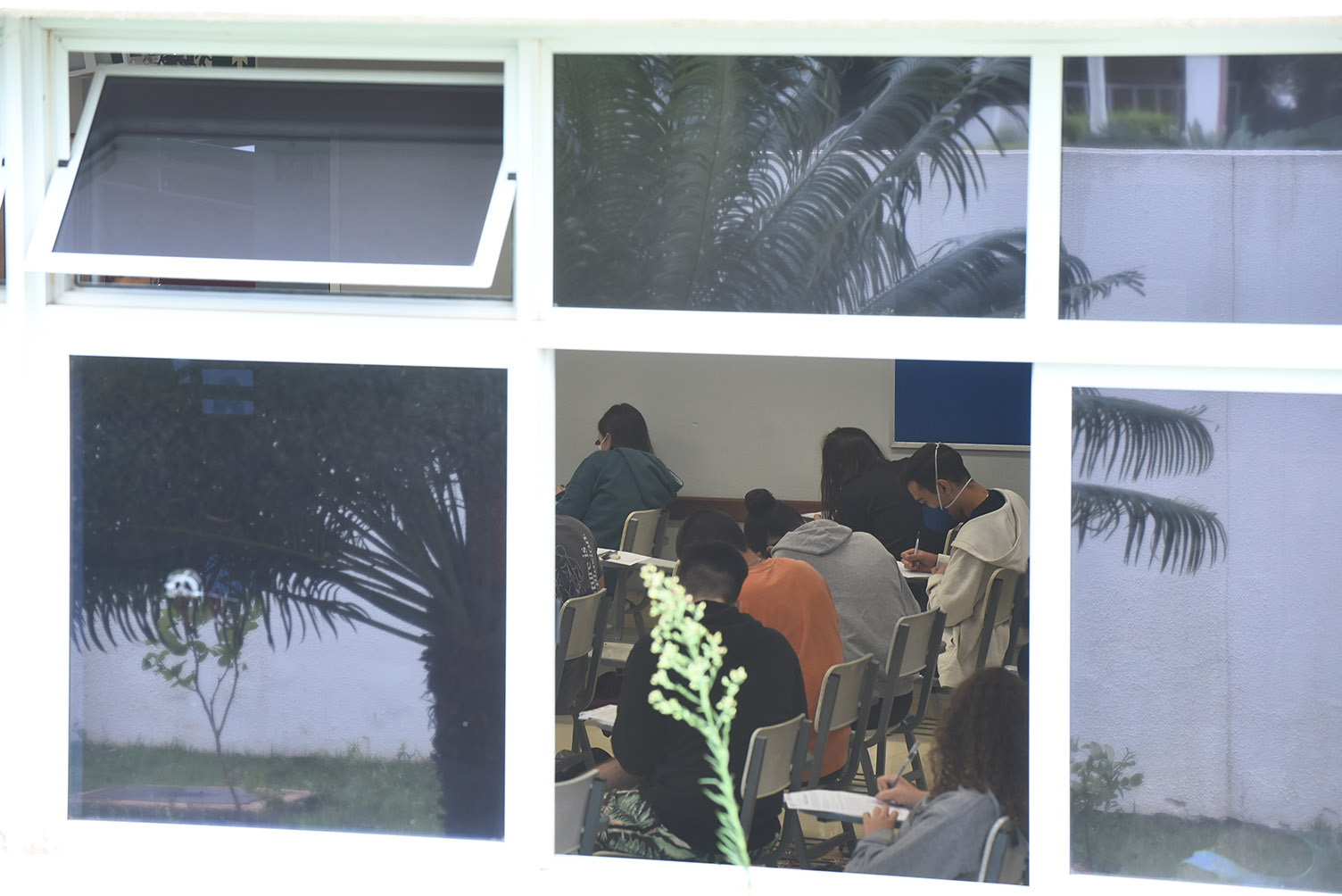 Candidatos durante provas da segunda fase do Vestibular Unicamp 2022, na PUC-Campinas Audiodescrição: Imagem realizada em área externa focada em uma janela de vidro de modo a permitir visualizar o interior de uma sala de aula, onde várias pessoas sentadas em carteiras com braço realizam prova. Há o reflexo de uma árvore no vidro da janela. Imagem 12 de 20.