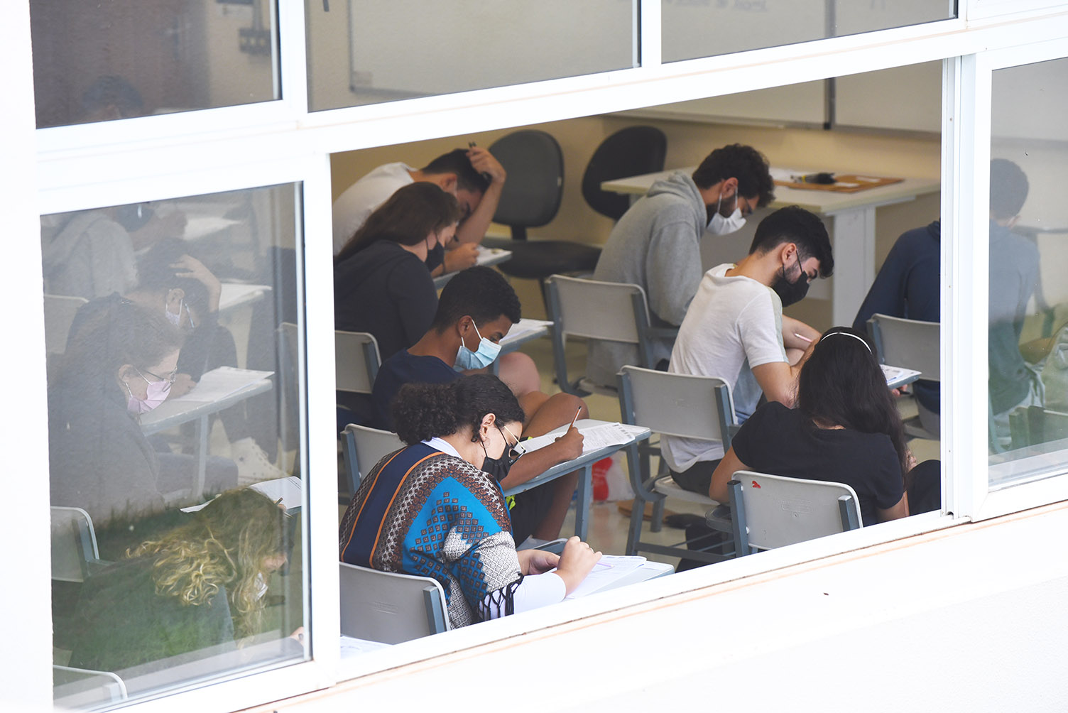 Candidatos durante provas da segunda fase do Vestibular Unicamp 2022, na PUC-Campinas Audiodescrição: Imagem realizada em área externa focada em uma janela de vidro de modo a permitir visualizar o interior de uma sala de aula, onde várias pessoas sentadas em carteiras com braço realizam prova. Imagem 13 de 20.