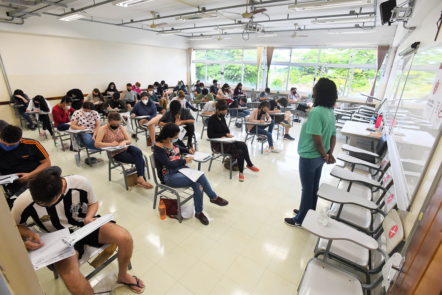 Candidatos durante provas da segunda fase do Vestibular Unicamp 2022, na PUC-Campinas Audiodescrição:Em sala de aula, imagem panorâmica e em perspectiva, cerca de 40 pessoas sentadas em carteiras escolares com braço, dispostas uma ao lado da outra, com cerca de 2 metros de distância, formando seis fileiras horizontais. Com olhares voltados para a direita da imagem, elas fazem prova de vestibular, segurando canetas e escrevendo em folhas de papel sulfite. À frente delas, ao centro, homem em pé, com as mãos unidas às costas, observa-os. Todos usam máscara contra covid-19. Imagem 16 de 20.