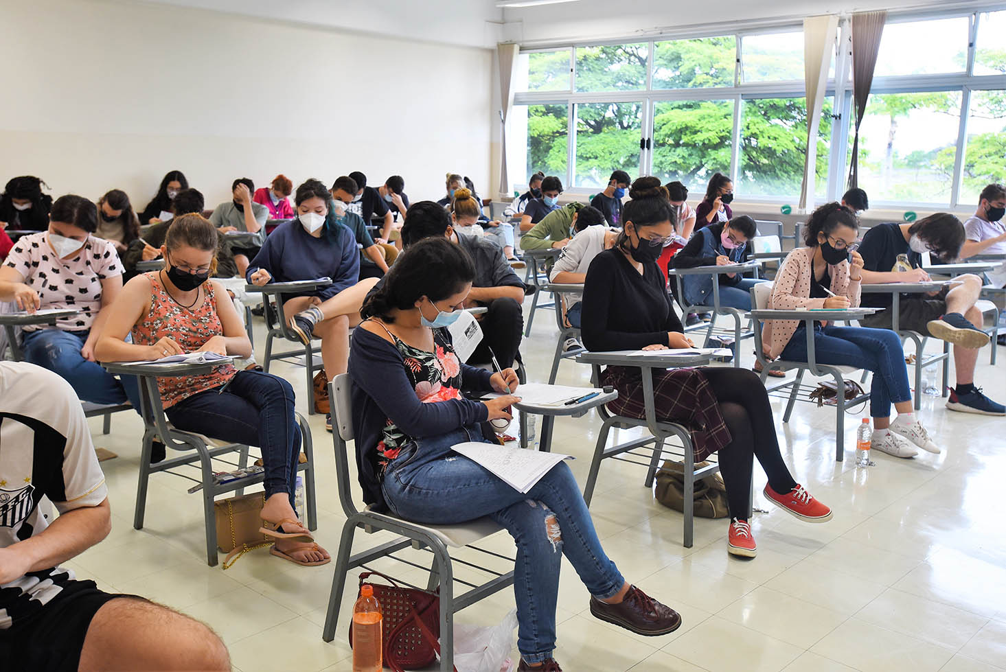 Candidatos durante provas da segunda fase do Vestibular Unicamp 2022, na PUC-Campinas Audiodescrição: Em ampla sala de aula, imagem panorâmica e em perspectiva, cerca de 40 pessoas sentadas em cadeira escolares com braço, dispostas uma ao lado da outra, com cerca de 2 metros de distanciamento, formando seis fileiras horizontais. Com olhares voltados para a direita da imagem, elas fazem prova de vestibular, segurando canetas e escrevendo em folhas de papel sulfite. Todos usam máscara de proteção contra covid-19. Imagem 17 de 20.