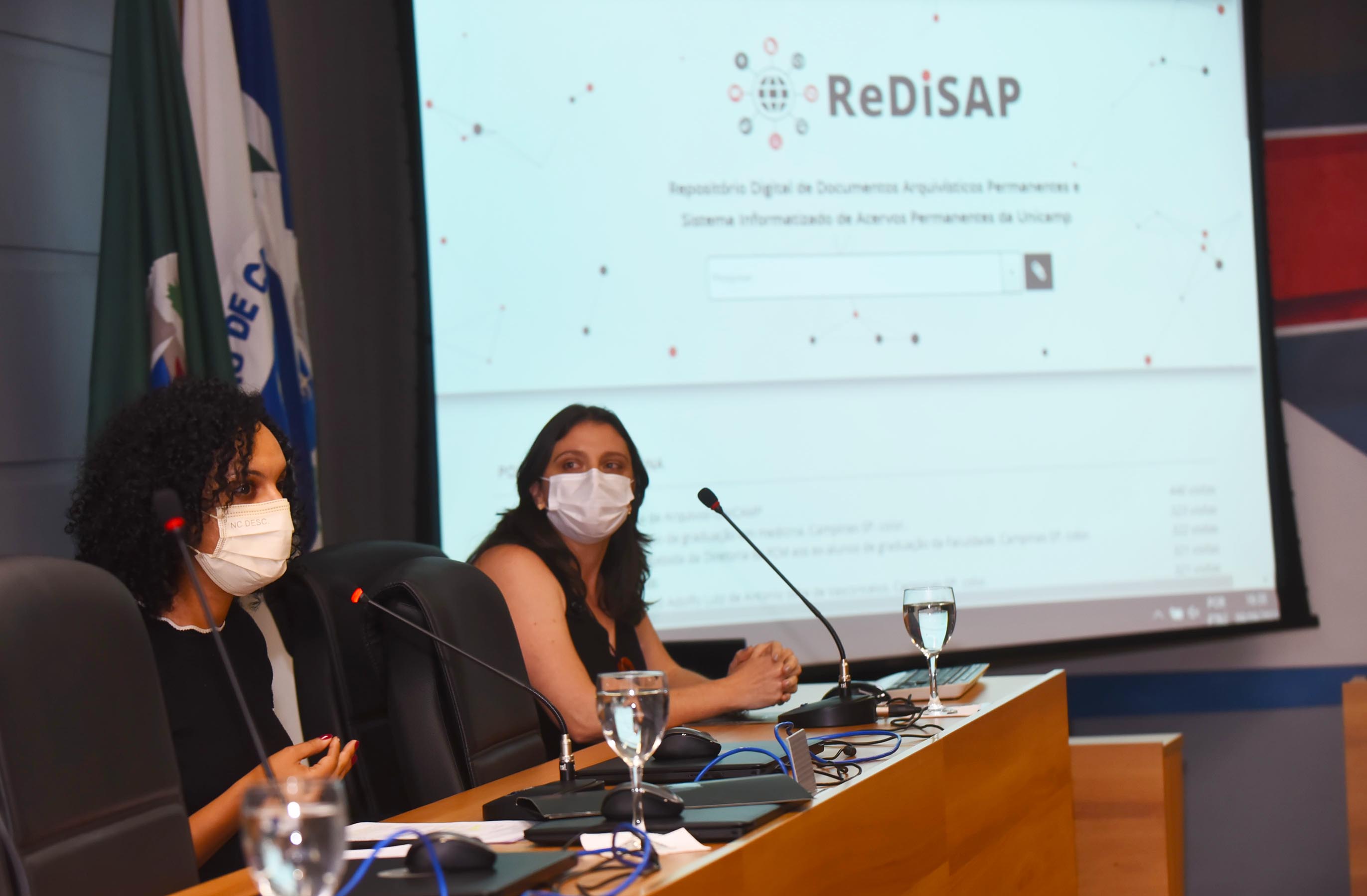 AD: no palco de um auditório, imagem em perspectiva e à curta distância de 2 mulheres sentadas em cadeiras, posicionadas uma ao lado da outra, à esquerda e voltadas para a direita da foto, sendo que a mulher em primeiro plano fala ao microfone de mesa, observada pela outra. À frente delas há uma extensa mesa de madeira, com taças de vidro com água e notebooks fechados. Ao fundo da imagem, tela de Data Show com fundo branco, ilustrações circulares, onde se lê em destaque Redisap. Imagem 2 de 6