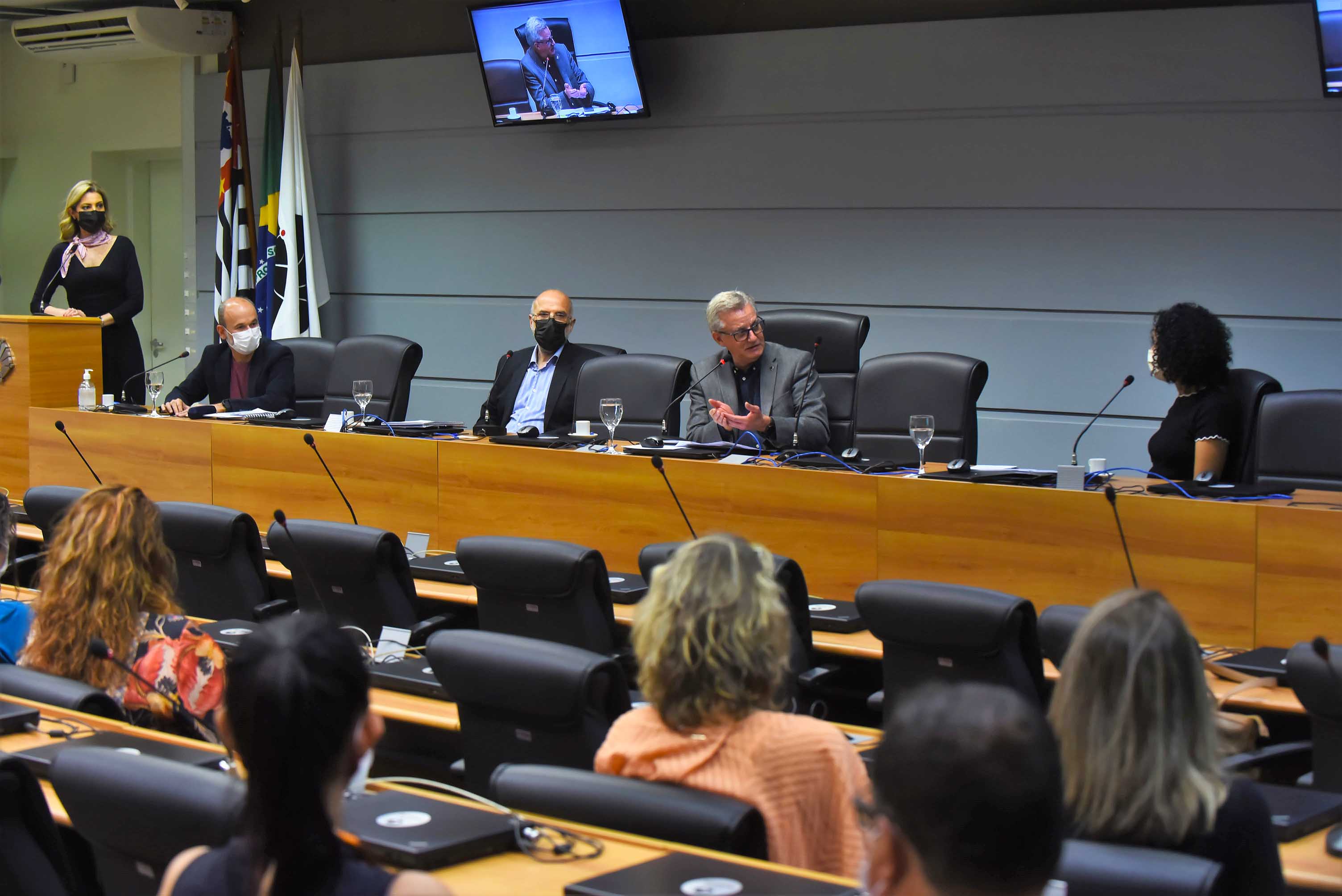 AD: em amplo auditório, imagem em perspectiva à média distância de 4 pessoas sentadas em cadeiras, uma ao lado da outra, voltadas à esquerda da foto, sendo que um homem, ao centro, fala ao microfone de mesa e gesticula com as mãos. Logo à frente, extensa mesa de madeira com 4 taças de vidro com água e notebooks fechados. Ao fundo da foto, à esquerda, 3 bandeiras afixadas em mastros e uma tv, ao alto. Mais à frente, de costas na imagem, o público do auditório, que está sentado em cadeiras. Imagem 3 de 6