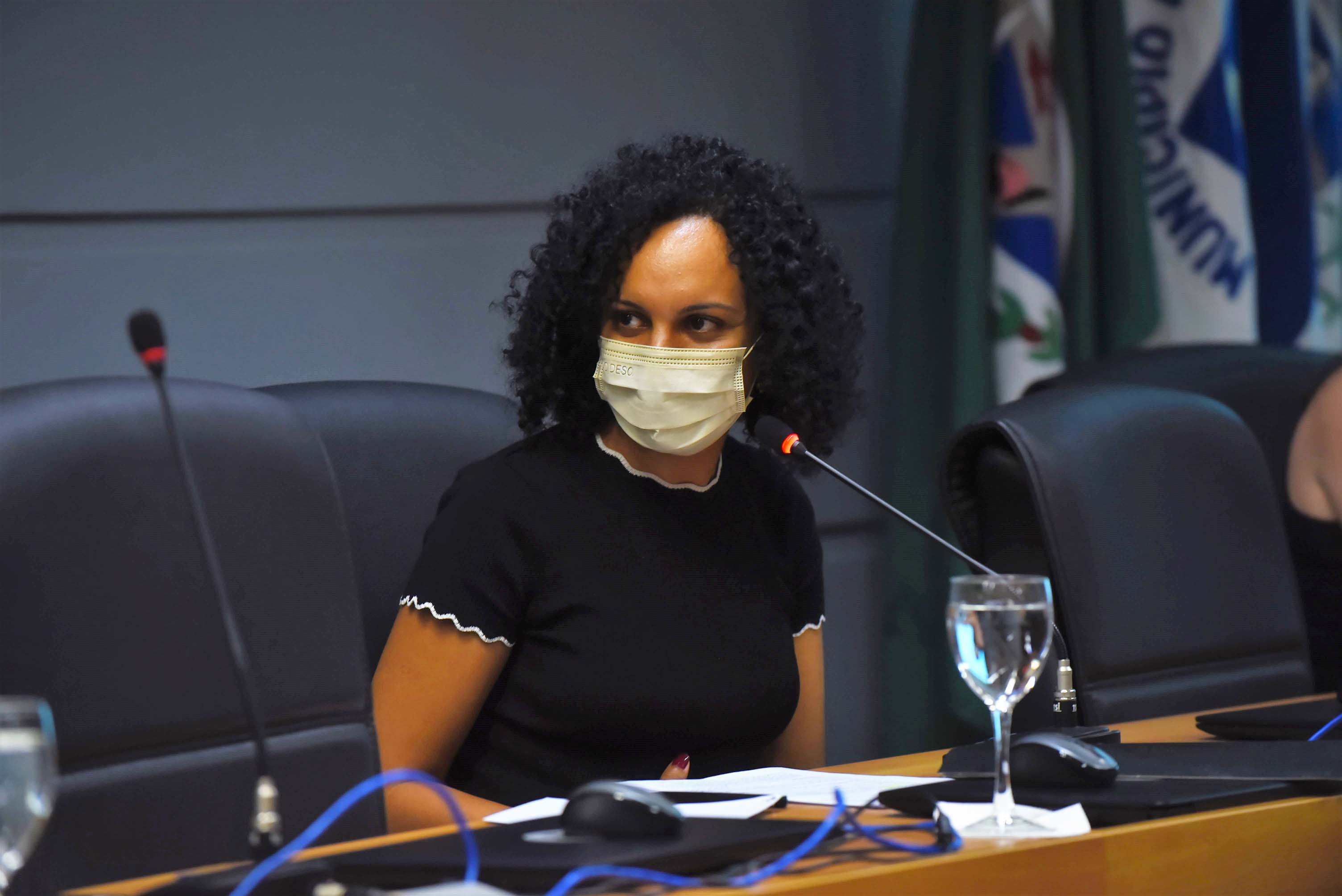 no palco de um auditório, imagem em perspectiva à curta distância de mulher sentada em cadeira, voltada para a esquerda da foto, sendo que ela fala ao microfone de mesa e mantém os braços baixados. À frente dela há uma extensa mesa de madeira 2 taças de vidro com água, notebooks fechados e mouses. Ela usa máscara de proteção respiratória e veste camiseta preta. Ao fundo, à esquerda, 3 bandeiras afixadas em mastros. Imagem 4 de 6