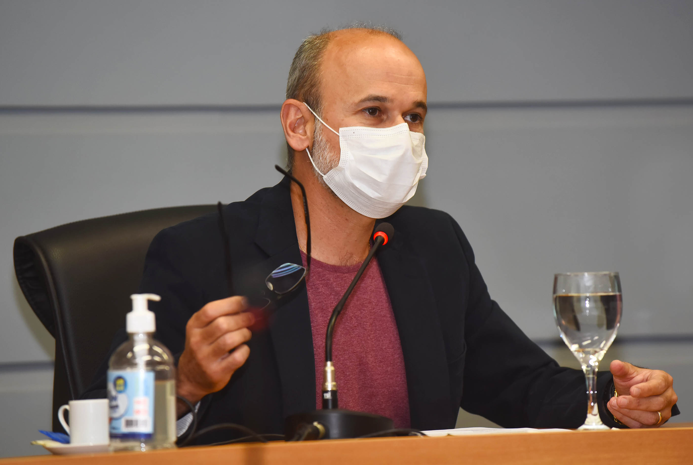 AD: no palco de um auditório, imagem em perspectiva à curta distância de homem sentado em cadeira, voltado para a direita da foto, sendo que ele fala ao microfone de mesa e mantém o braço esquerdo apoiado sobre uma mesa de madeira, posicionada À frente, e segura os óculos a mão direita. Sobre a mesa há uma taça de vidro com água, xícara e tubo de álcool gel. Ele usa máscara de proteção respiratória e veste terno preto. Ao fundo, parede com frisos horizontais em baixo relevo. Imagem 5 de 6