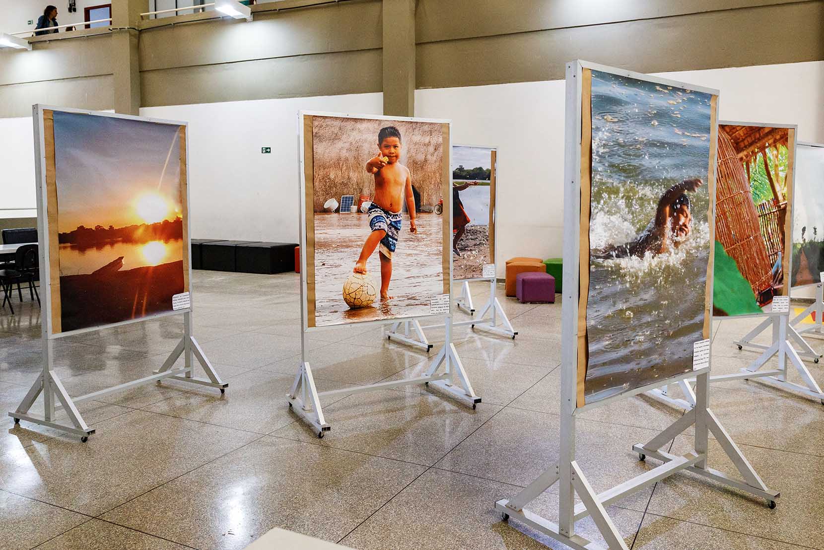 Exposição fotográfica “A diversidade cultural dos alunos e alunas indígenas do Instituto de Geociências (IG) – Unicamp”: fotos retratam as comunidades de onde vieram estudantes indígenas hoje presentes na unidade Audiodescrição (AD). Foto: Felipe Bezerra. Em um amplo saguão, imagem em perspectiva à média distância de 6 fotos coloridas, em tamanhos de cerca de 90 centímetros por 70 centímetros que se encontram afixadas em expositores verticais de madeira, e compõem uma exposição fotográfica. As fotos trazem temática indígena como: menino que nada em rio e menino em pé, com o pé direito sobre uma bola, tendo ao fundo uma ampla maloca, e que aponta para frente com o indicador direito. Imagem 1 de 6
