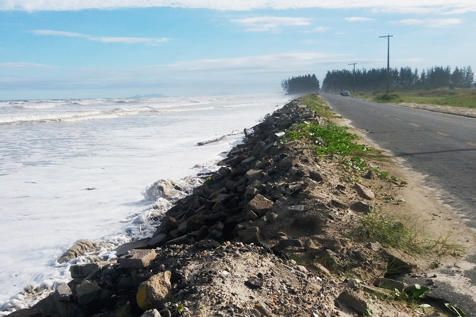 foto mostra um trecho da costa do litoral sul, onde as águas do mar batem contra pedras bem próximas a uma avenida beira-mar
