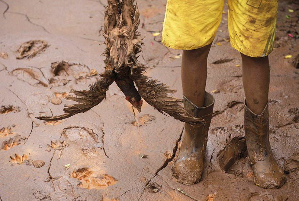 Foto de mostra os pés de uma criança enterrados na lama. Ele usa botas, bermuda branca e está segurando uma galinha que aparece de cabeça para baixo.