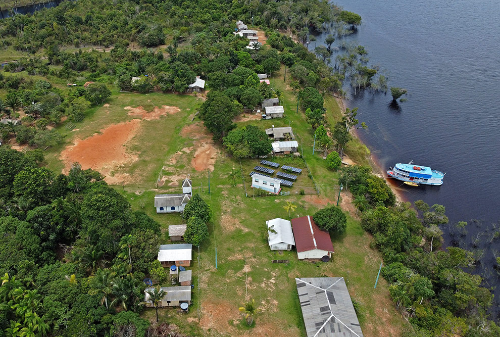 Foto aérea que mostra um conjunto de casas espalhadas entre árvores na beira de um rio. Na margem do rio há um barco.