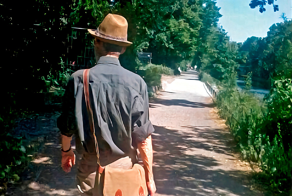 Foto de um homem que aparece da cintura para cima. Ele está de costas, está usando uma camisa escura e chapéu. Ele está caminhando em um jardim.