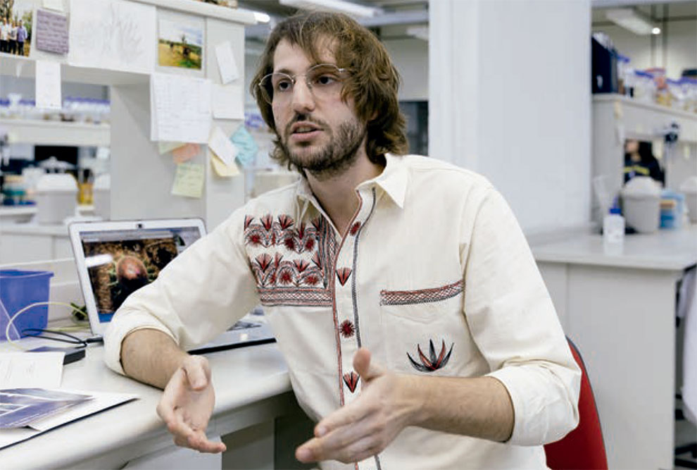 Foto de um homem que está sentado em um laboratório. Ele é branco, tem cabelos longos, ondulados, barba e bigode. Está usando camisa branca com desenhos de plantas.