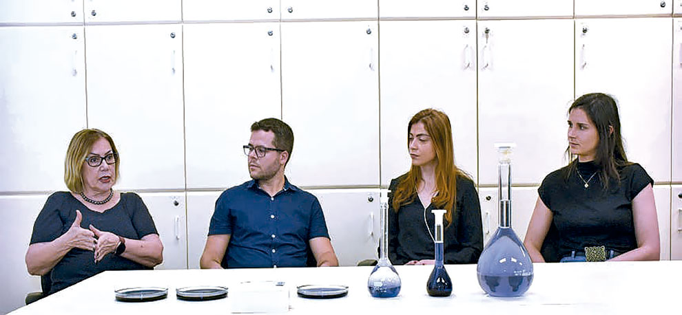 Foto mostra quatro pessoas atrás de uma mesa. Na mesa há amostras de líquidos em vidros de formas e tamanhos variados.