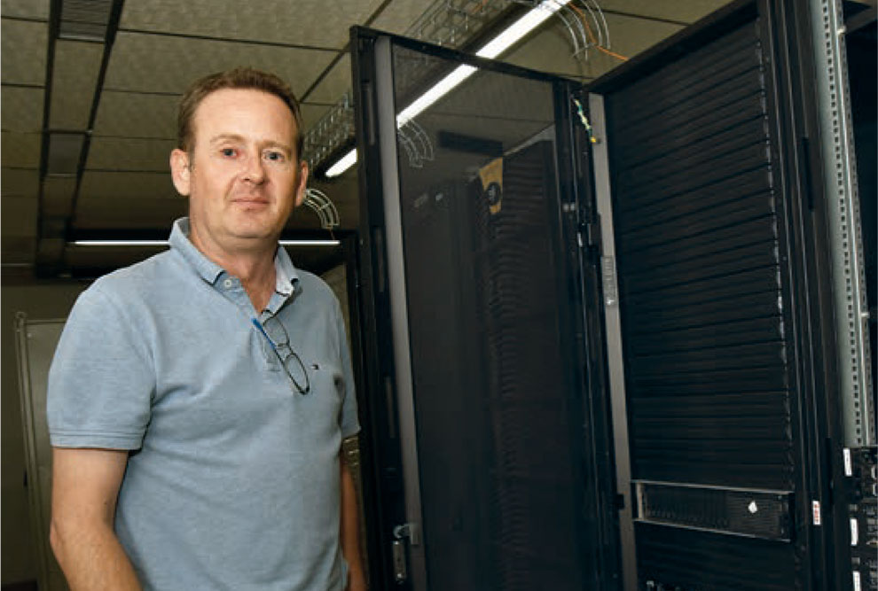 Foto de um homem que está de pé, ao lado de um servidor de computador. Ele é branco, tem o cabelo curto, está usando camiseta polo e calça jeans.